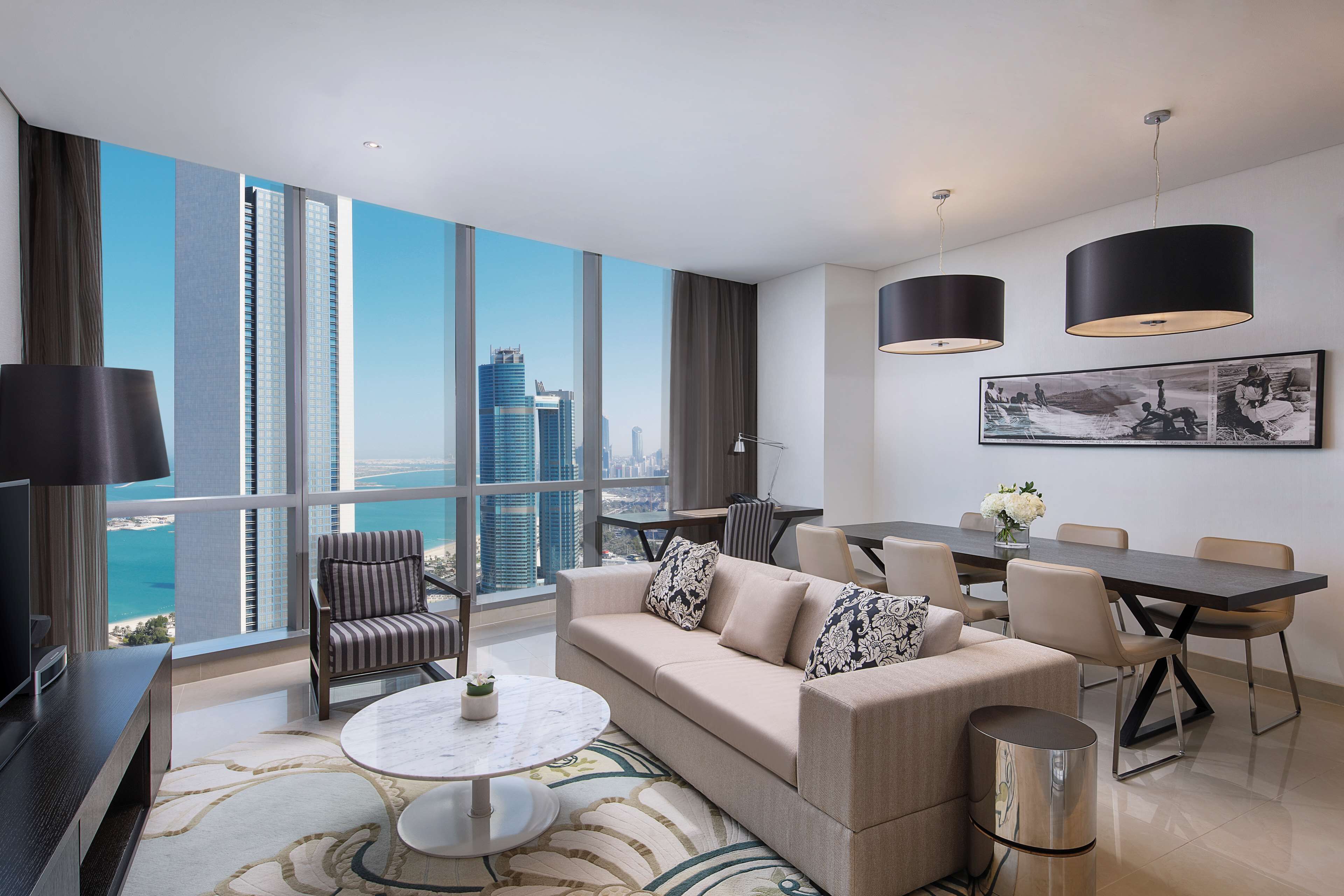 Una sala de estar moderna con sofá, mesa de centro y comedor. Amplias ventanas ofrecen vistas a la ciudad.