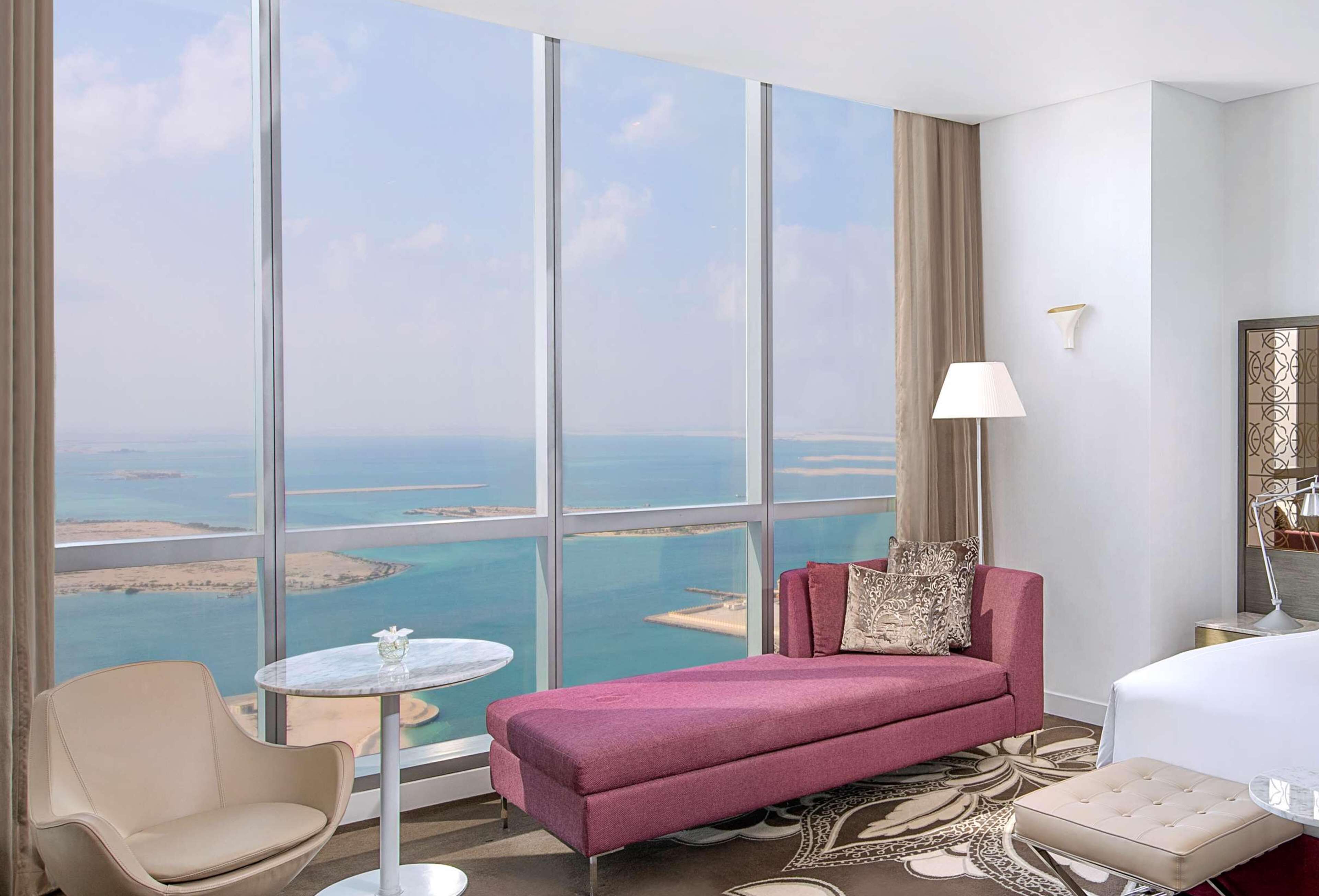 Habitación de hotel con un amplio ventanal que ofrece vistas de la costa, un sofá rosa, una mesa redonda y una cama.