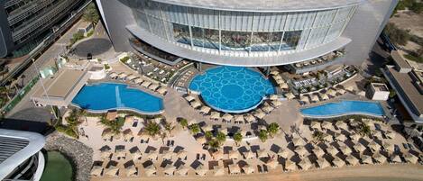 A modern hotel with multiple swimming pools, sun loungers, and a beachfront view.