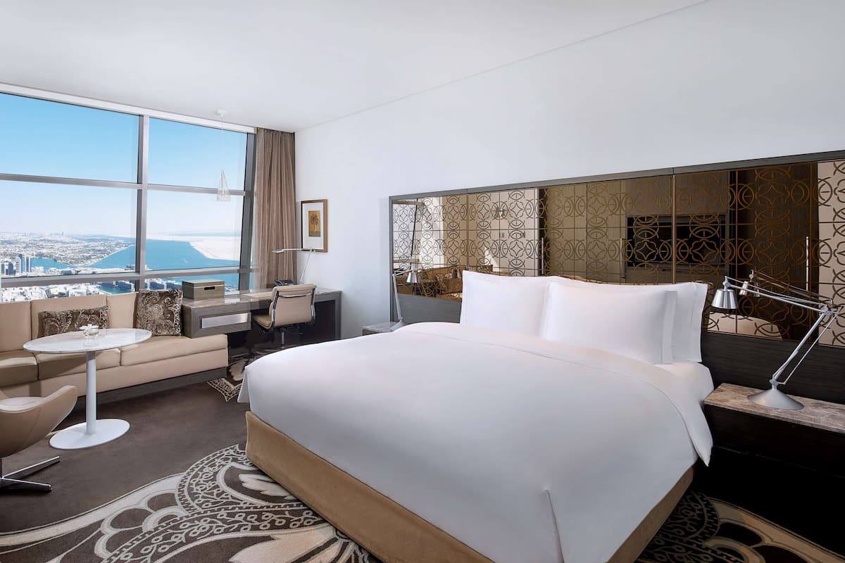 Habitación de hotel con una cama grande, un escritorio, una silla, una mesita y un amplio ventanal con vistas a la ciudad.