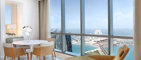 A modern hotel room with a large panoramic window, a round dining table with chairs, a sofa, and a vase with flowers.