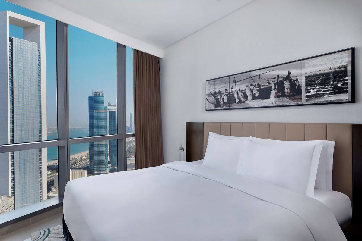 Una habitación de hotel moderna con una cama grande, vistas a la ciudad y una fotografía histórica en un marco en la pared.