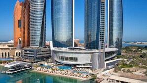 Un hotel moderno con piscina frente al agua, varios edificios altos y un puerto deportivo.