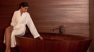 Une personne vêtue d’un peignoir blanc est assise au bord d’une baignoire en bois, dans un cadre de spa.