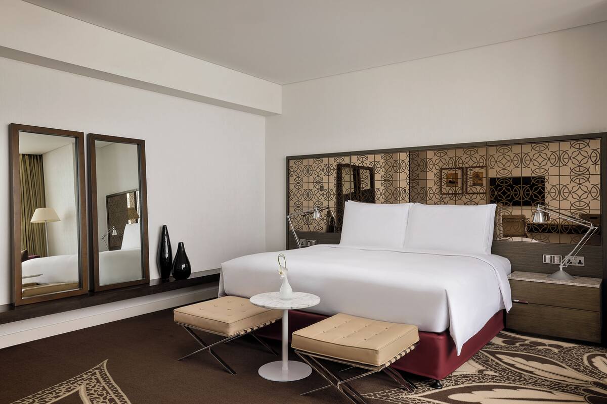 Una habitación de hotel moderna con una cama grande, una mesita redonda pequeña, dos sillas, una mesita de noche con una lámpara y un panel decorativo en la pared.