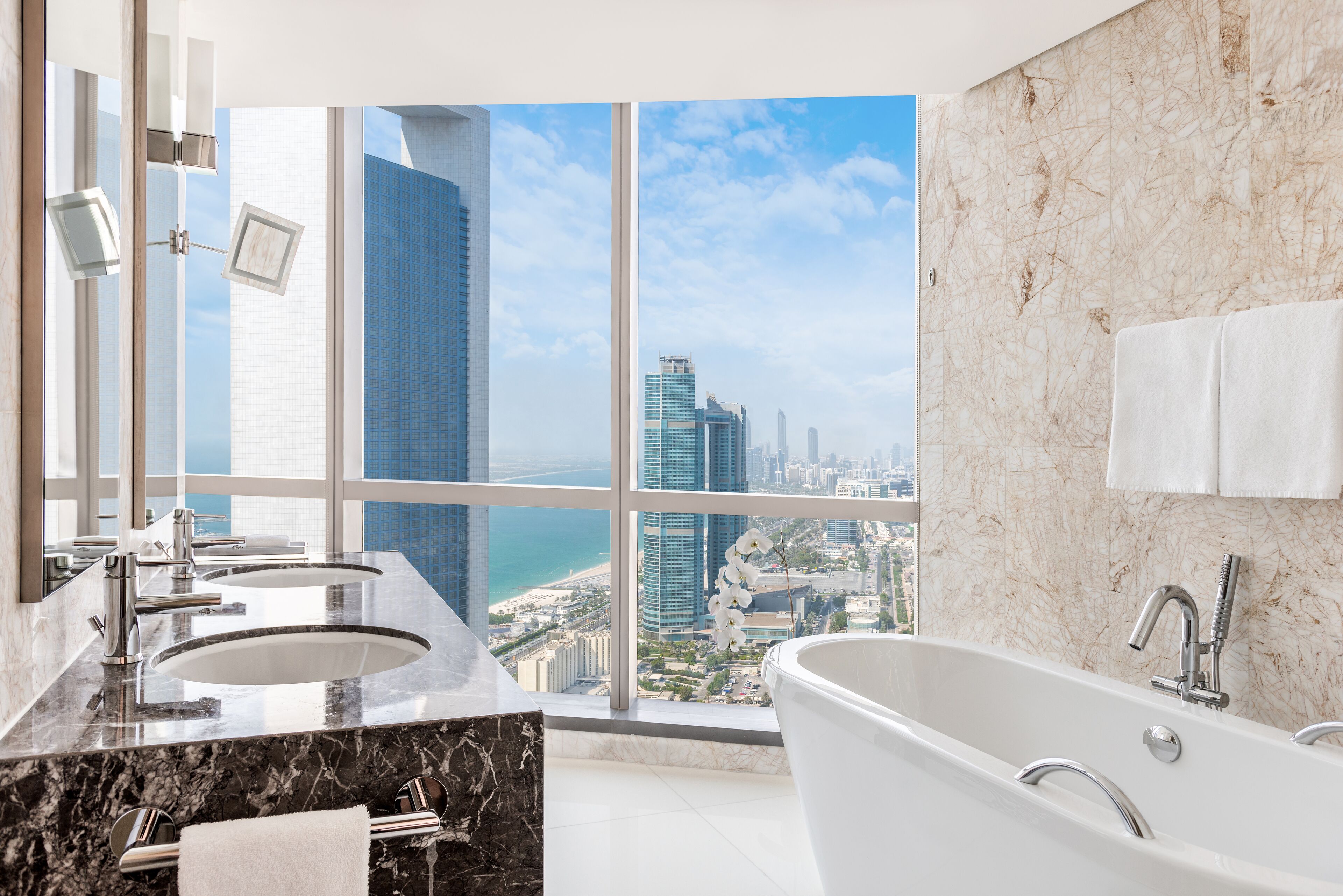 A modern bathroom with dual sinks, a large bathtub, and a city view through floor-to-ceiling windows.