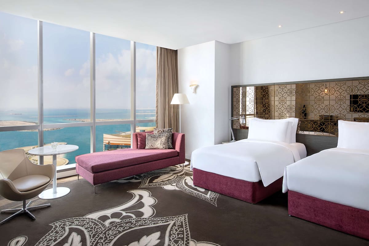 Habitación de hotel con un ventanal grande, un sofá, dos camas, una mesita y una lámpara.