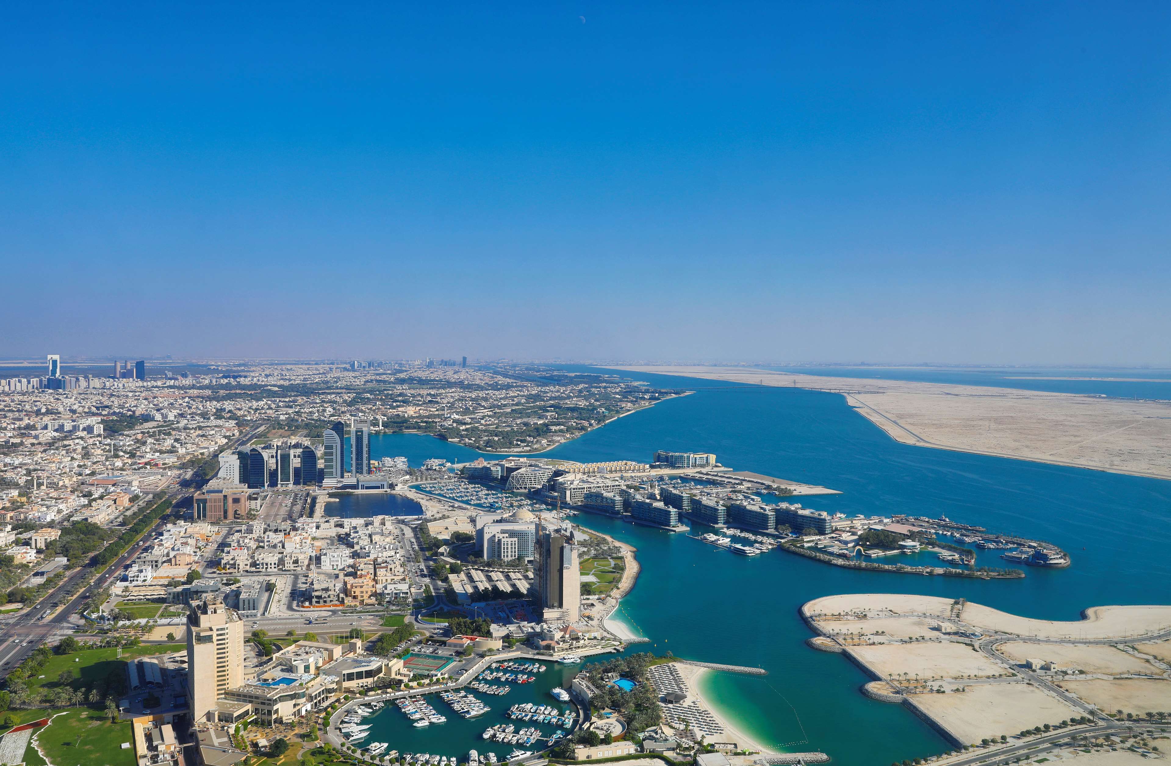 An aerial view of a coastal city with a marina, high-rise buildings, and a clear blue sky.