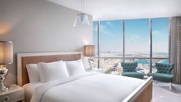 A hotel room with a large bed, two bedside lamps, a view of a cityscape through large windows, and two upholstered chairs.