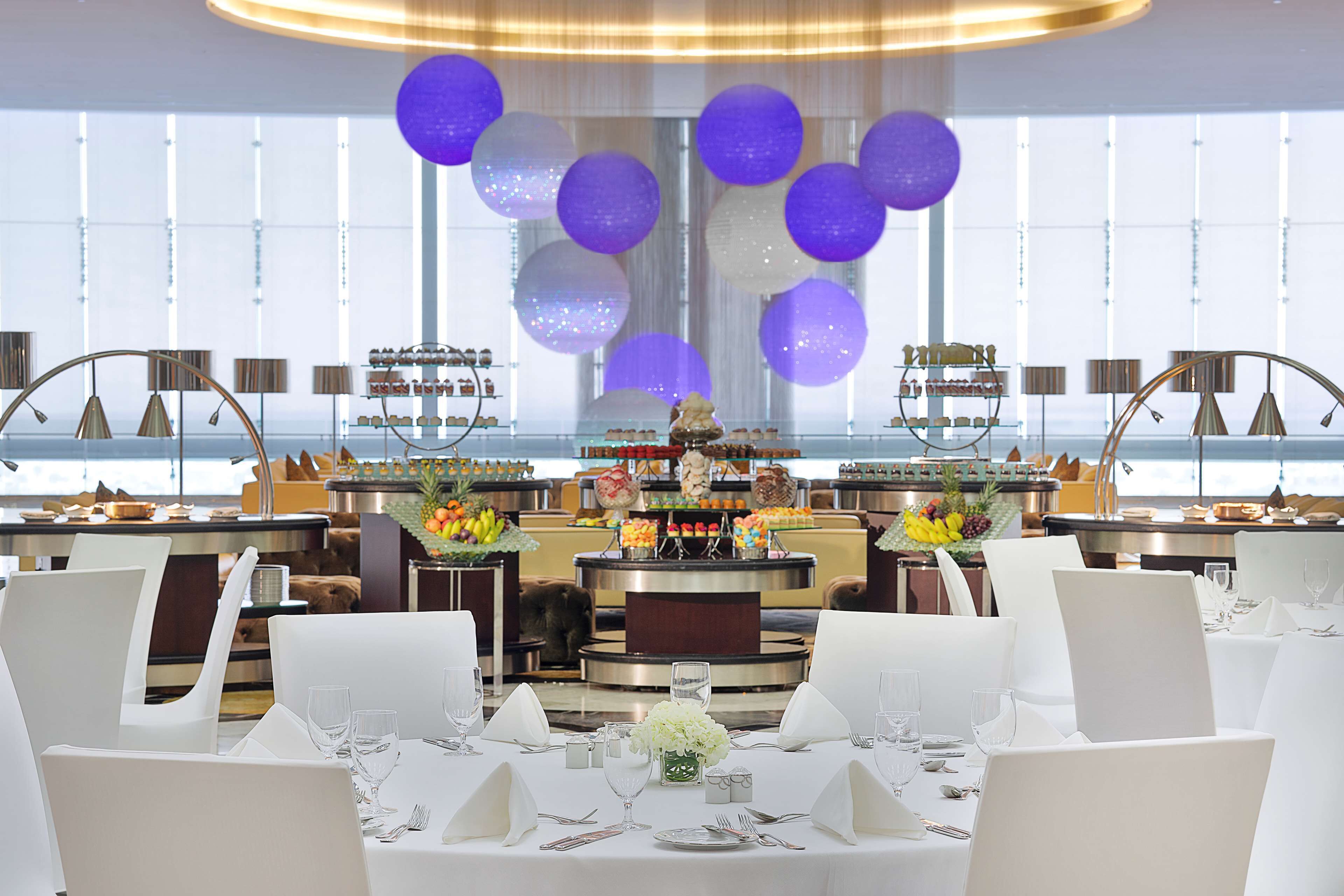 A restaurant with white chairs, round tables, and a central buffet area.