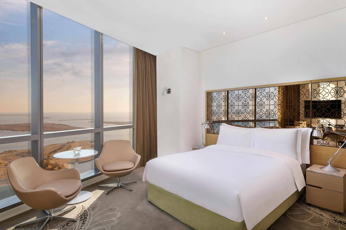 Habitación de hotel con una cama grande, dos sillas, una mesita y vistas al paisaje a través de amplias ventanas.