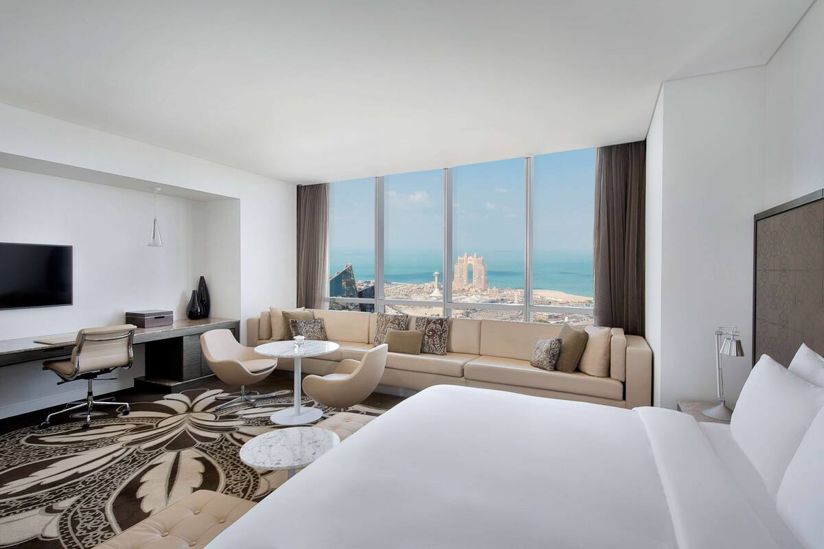 Habitación de hotel con una cama grande, un sofá, un escritorio con una silla, un televisor y vistas al mar y al paisaje urbano.