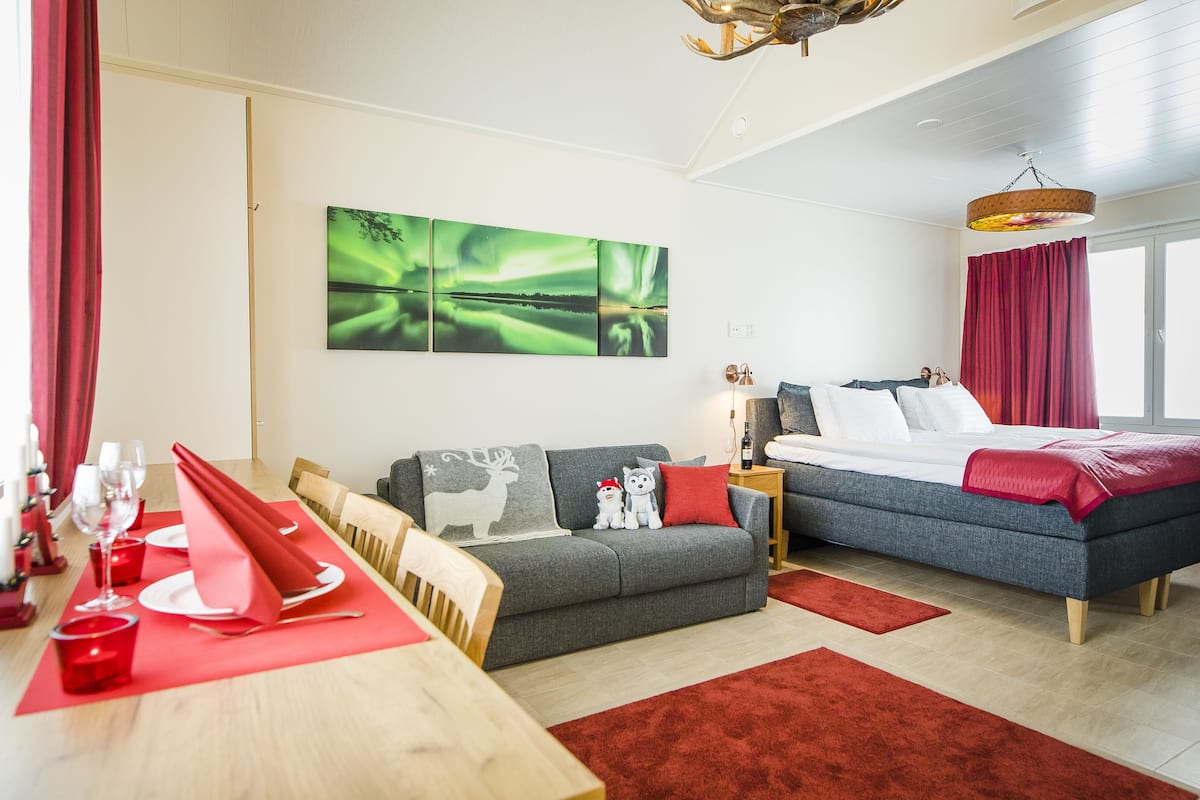 A modern hotel room with a dining area, a grey sofa, a bed with red bedding, and a large window.