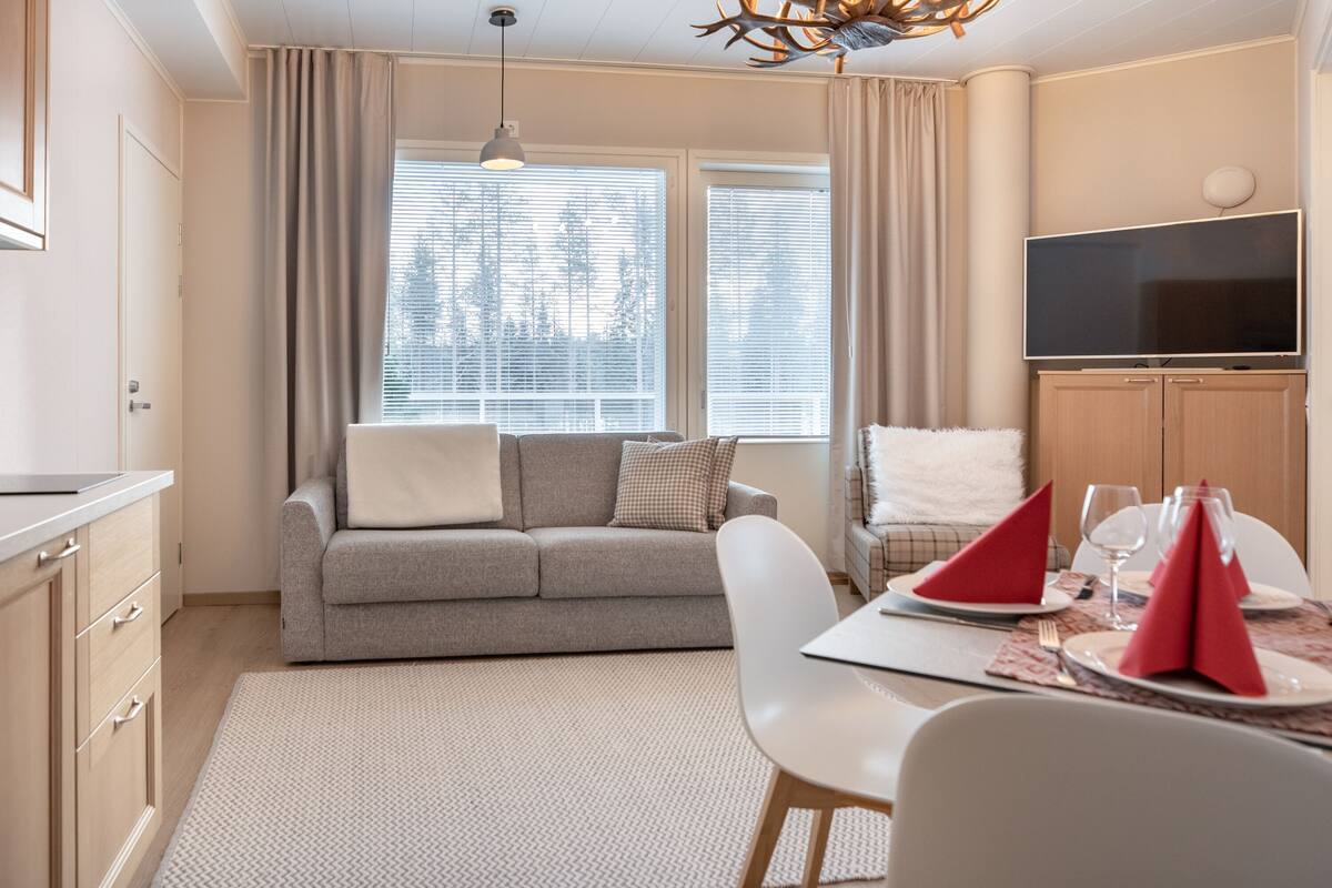 A modern living room with a sofa, a dining table set for two, and a flat-screen TV mounted on the wall.