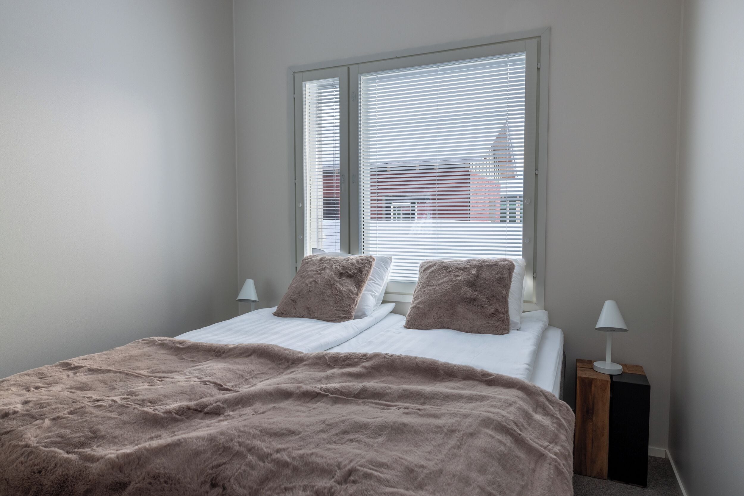A bedroom with a bed, two pillows, a window with blinds, and a bedside lamp.