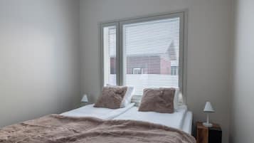 A bedroom with a bed, two pillows, a window with blinds, and a bedside lamp.
