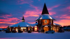 A wooden building with a steeply pitched roof and a prominent tower, surrounded by snow and parked cars. - Santa Claus Holiday Village (Rovaniemi)