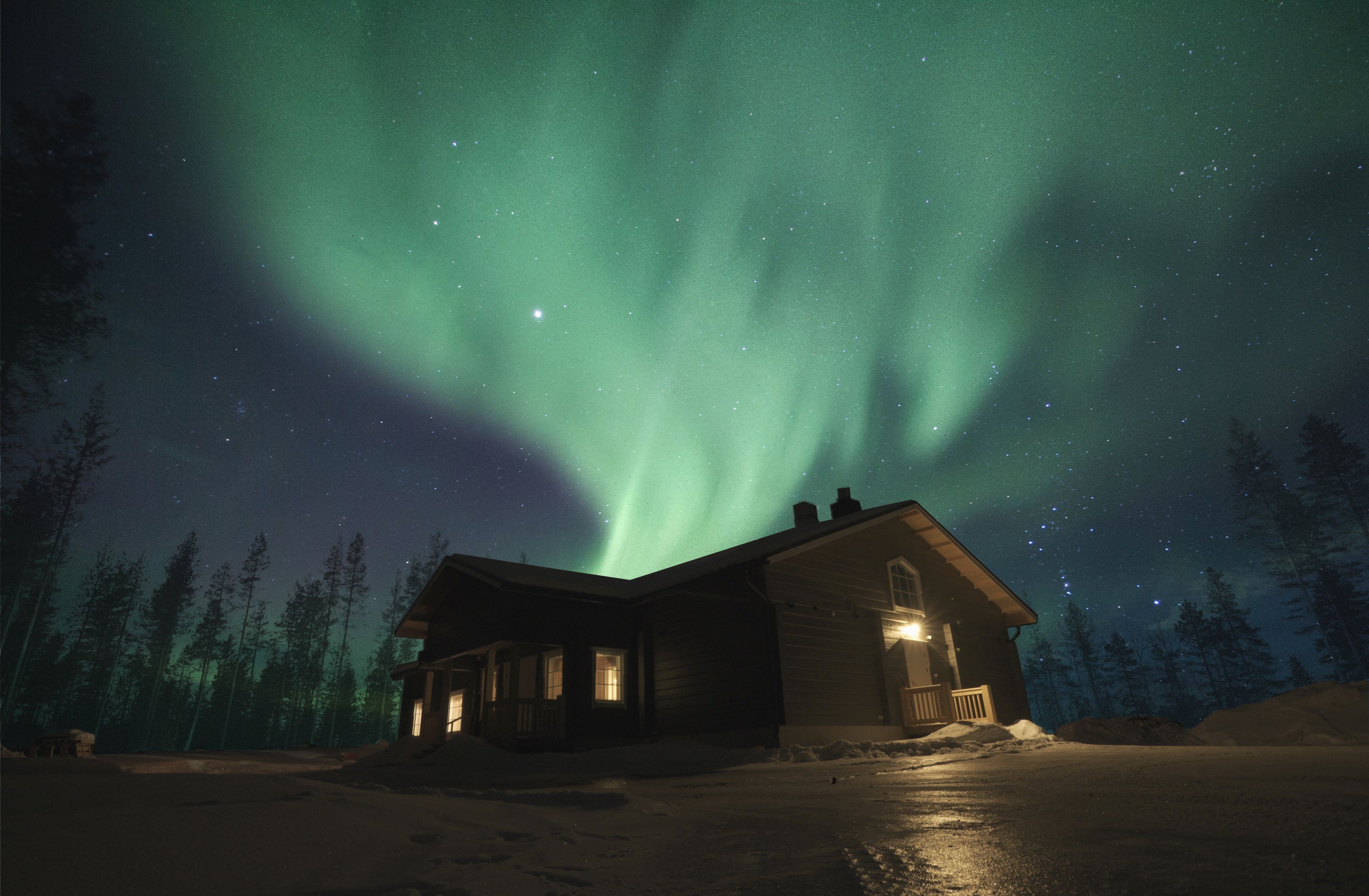 A cabin with a snow-covered ground, surrounded by trees, under a night sky with visible stars and the aurora borealis.