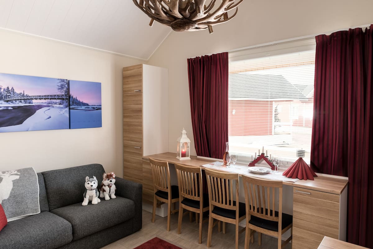 A cozy room with a grey sofa, two plush dog toys, a wooden dining table with four chairs, a red curtain, and a window with blinds.