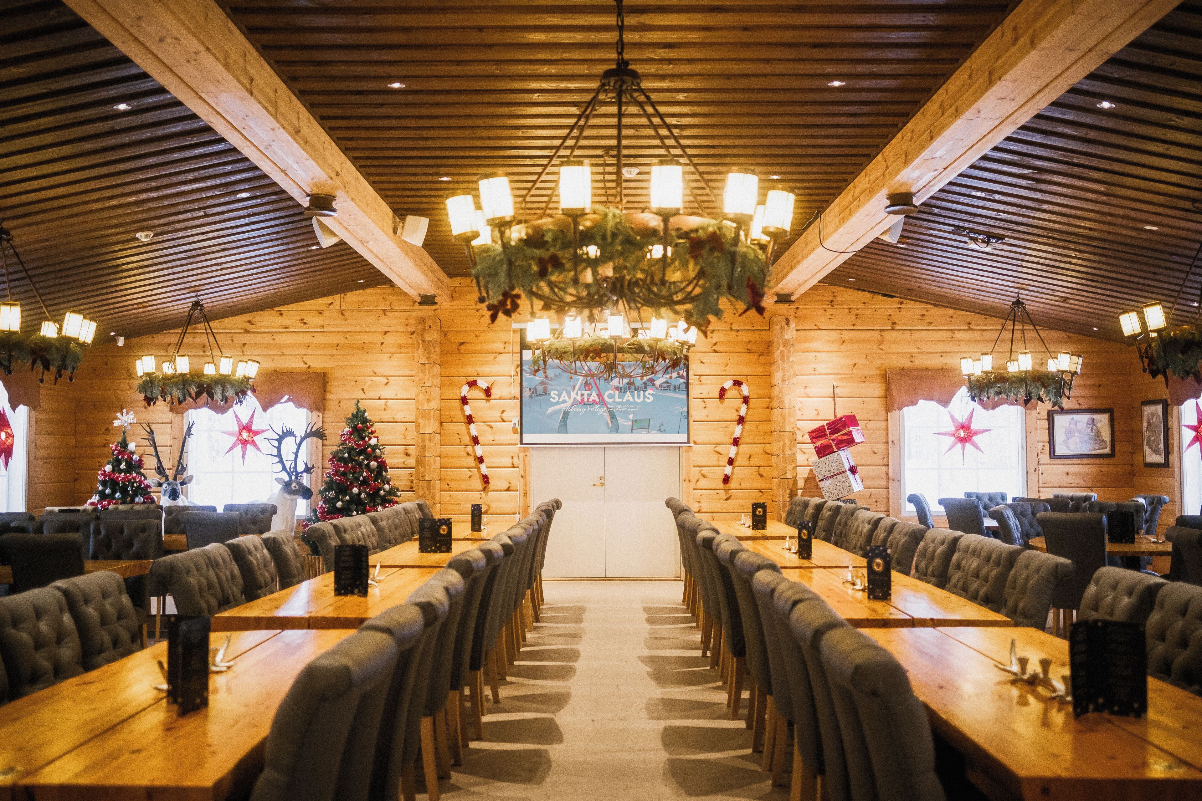 Een restaurant met lange houten tafels en stoelen, een kroonluchter, kerstversiering, en een groot scherm waarop 'SANTA CLAUS' staat.