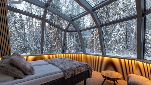 A bedroom with a bed, a chair, and a view of snow-covered trees through large windows. - Santa Claus Holiday Village (Rovaniemi)