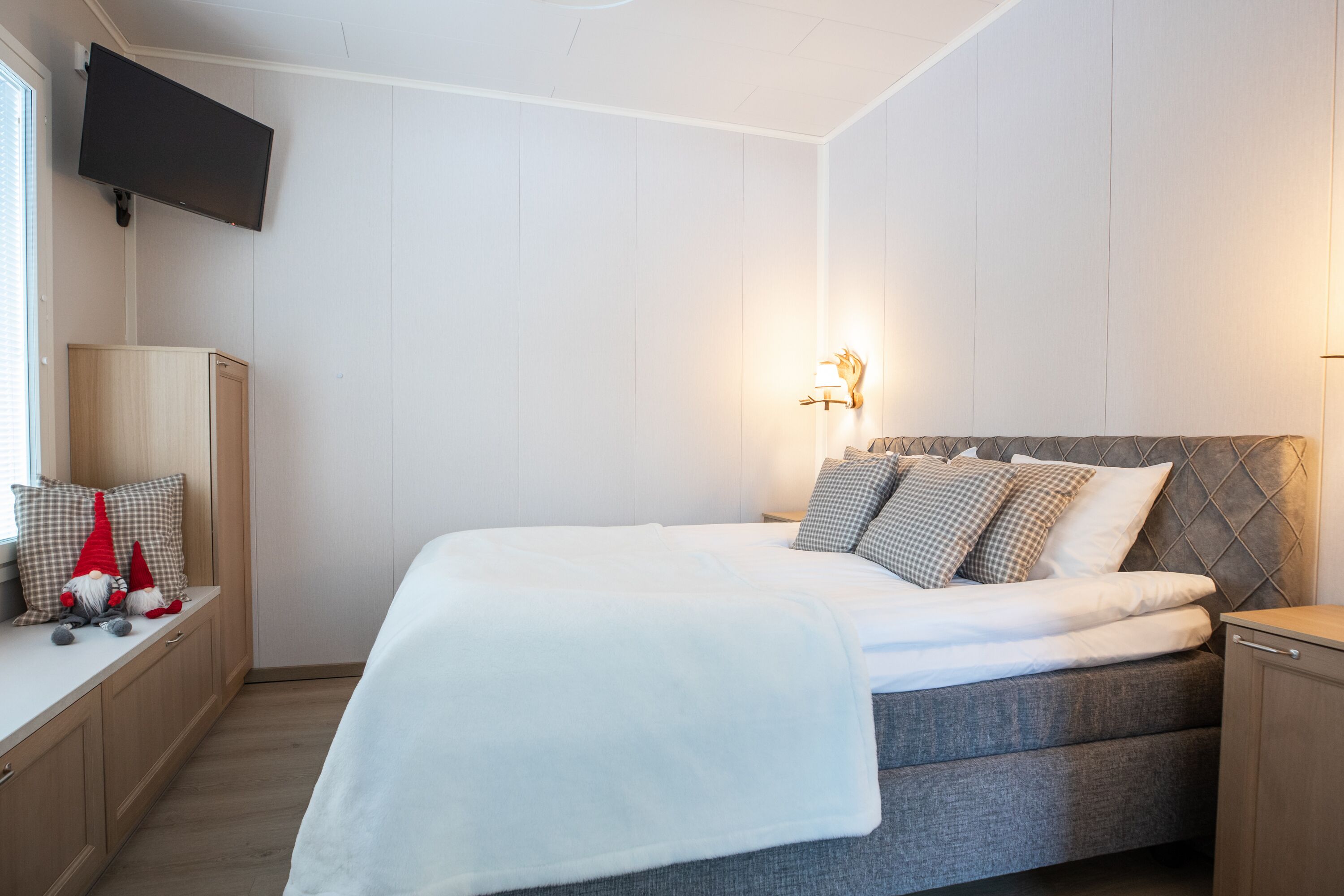Ein ordentlich eingerichtetes Schlafzimmer mit einem Bett, Nachttischen, einem an der Wand befestigten Fernseher und einer Wandleuchte.