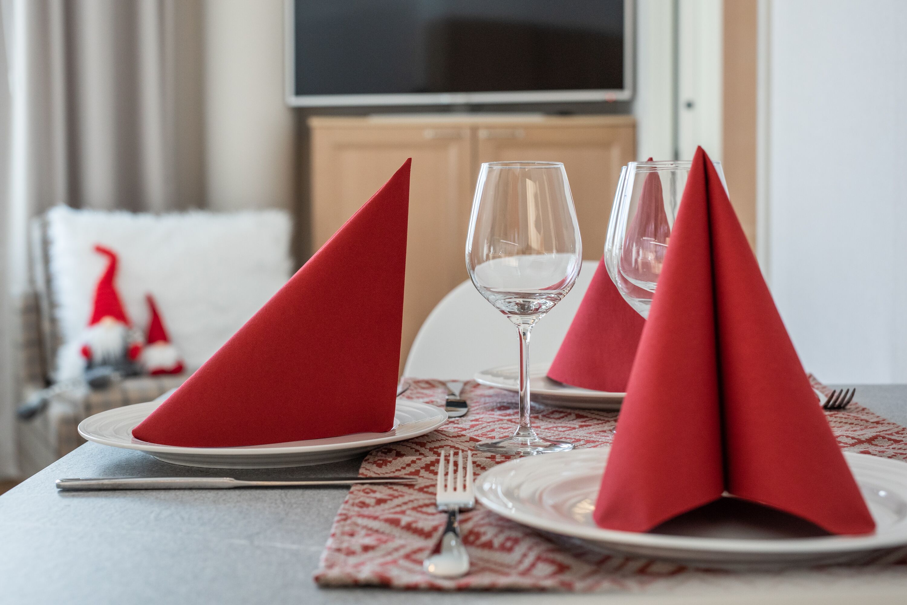 A dining table set with red napkins, wine glasses, and plates, with a patterned placemat and a fork on the table.