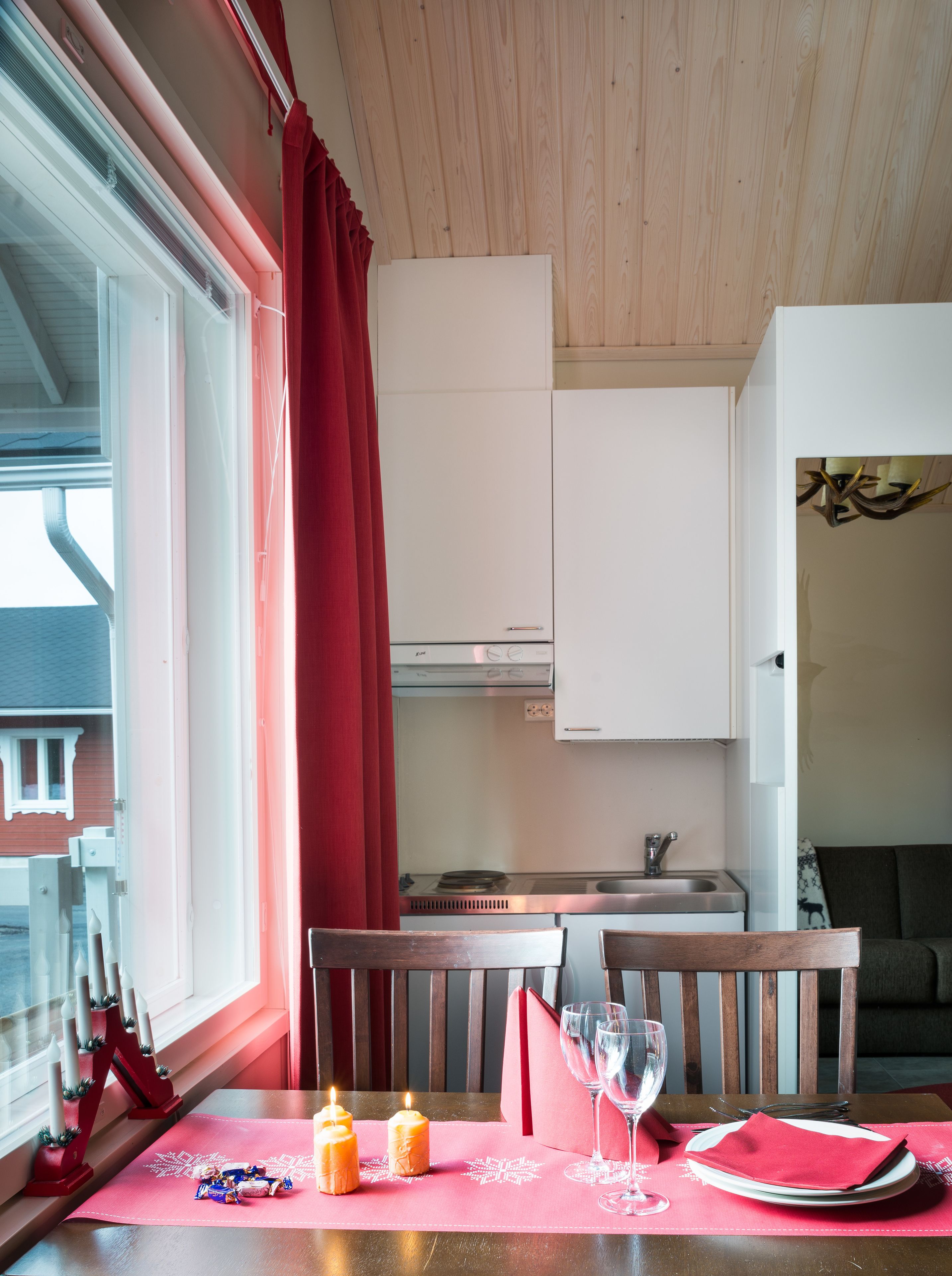 A compact kitchen with white cabinets, a dining area with a table set for two, and a window with red curtains.