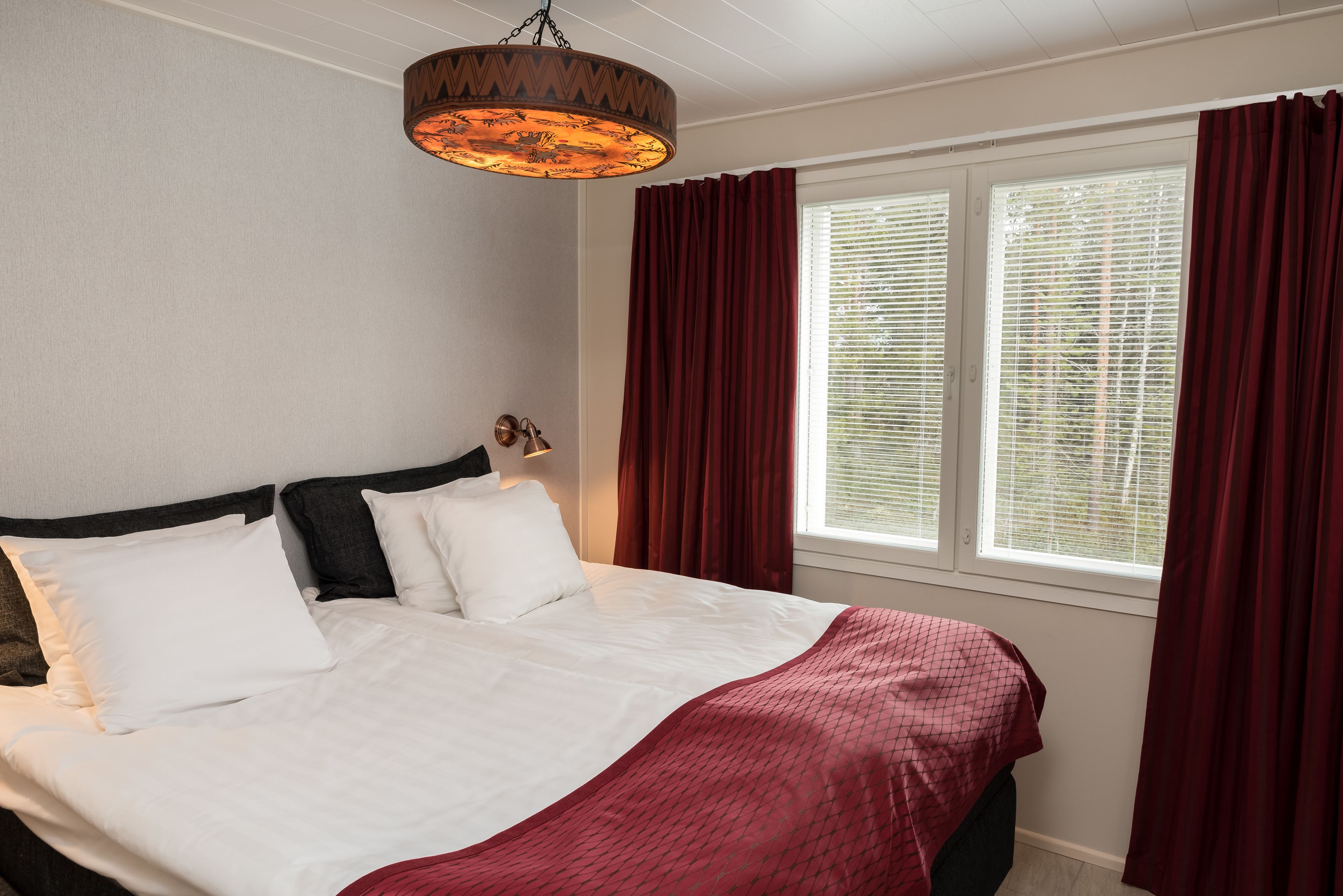 A neatly made bed with white and red bedding, a hanging lamp, and a window with red curtains.