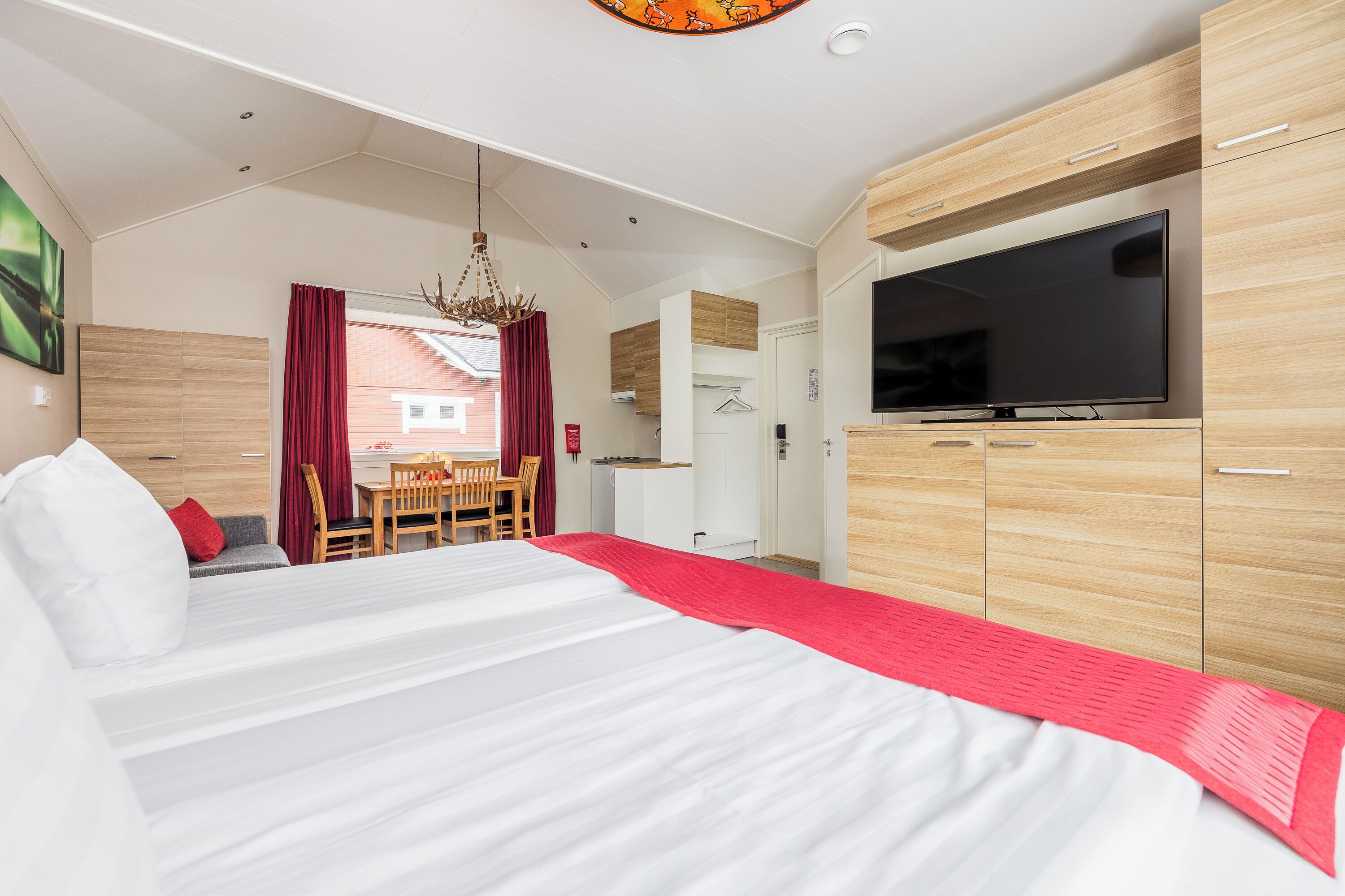 A modern hotel room with a large bed, a TV, wooden cabinets, and a dining area with a table and chairs.