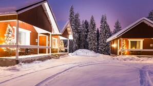 A snow-covered landscape with two wooden cabins, each with a lit Christmas tree and a number sign indicating room numbers. - Santa Claus Holiday Village (Rovaniemi)
