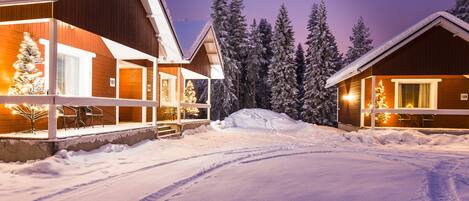 A snow-covered landscape with two wooden cabins, each with a lit Christmas tree and a number sign indicating room numbers.