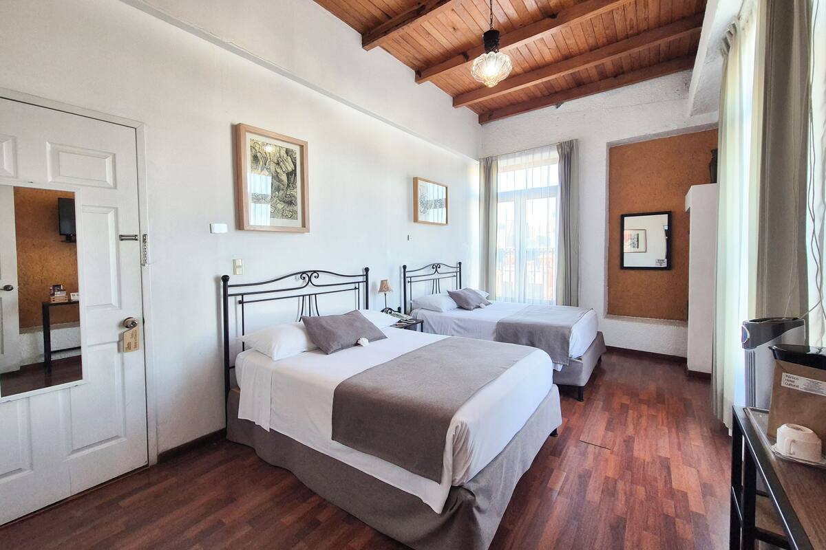 Une chambre d’hôtel avec deux lits, un plancher en bois, un plafond avec des poutres apparentes et une fenêtre avec des rideaux.