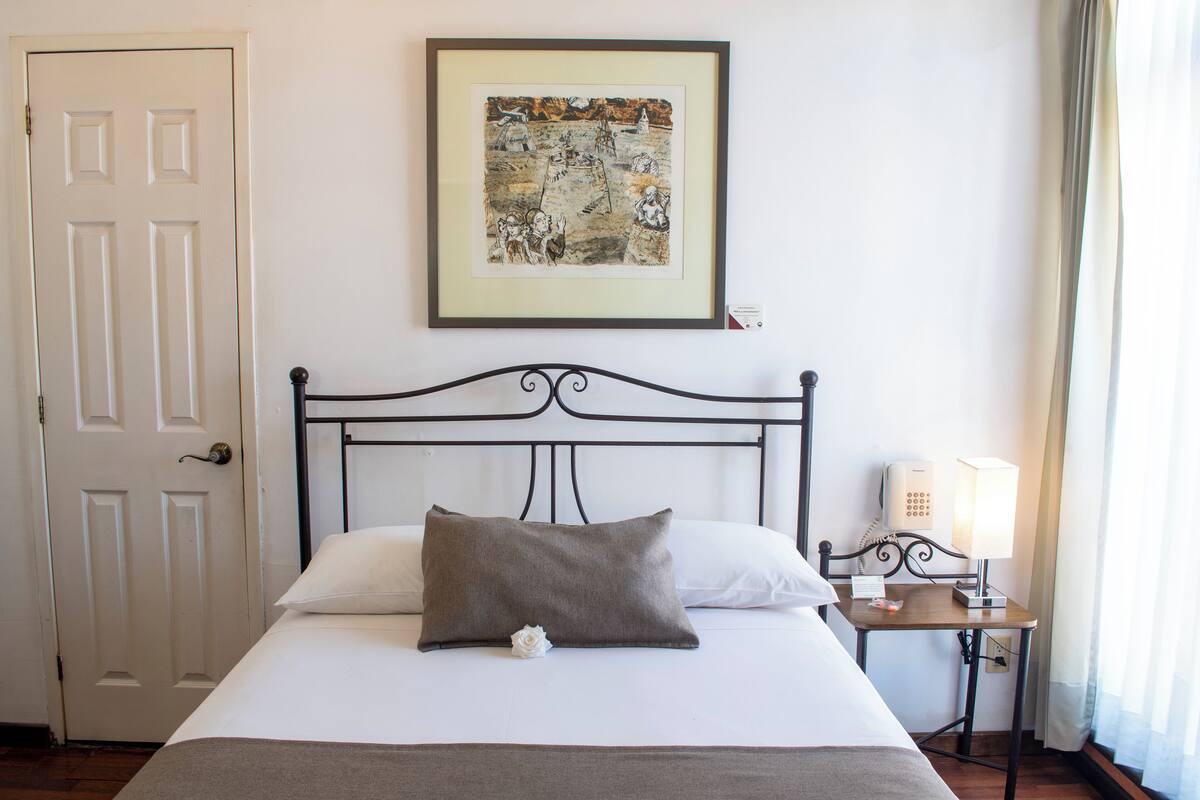 Un lit bien fait, avec un oreiller et une seule rose blanche, une œuvre d’art encadrée au-dessus du lit, une table de chevet avec une lampe et une porte sur le côté gauche.