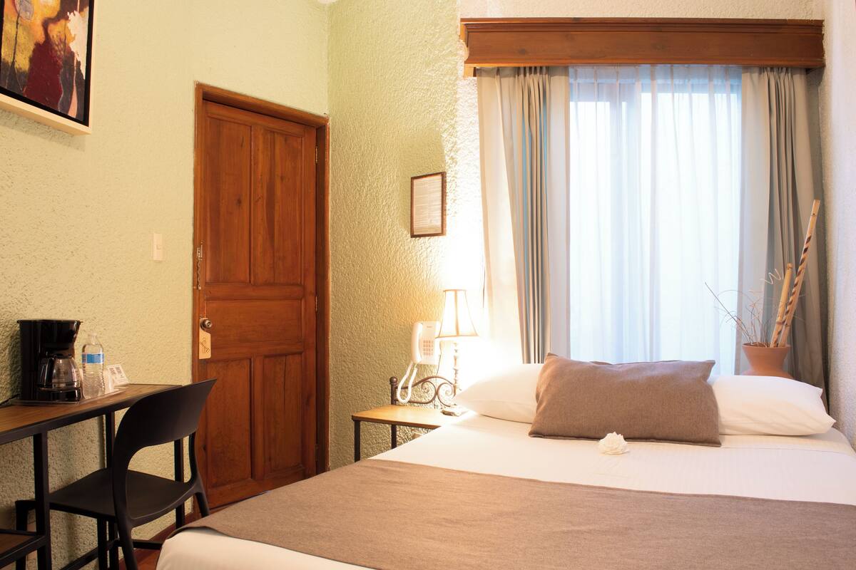 Une chambre d’hôtel comprenant un lit, un bureau avec une chaise, un téléphone et une fenêtre avec des rideaux.