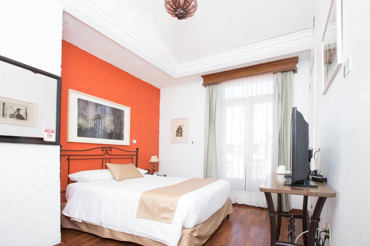 Une chambre d’hôtel équipée d’un lit, d’un bureau, d’une télévision et de tableaux encadrés aux murs.
