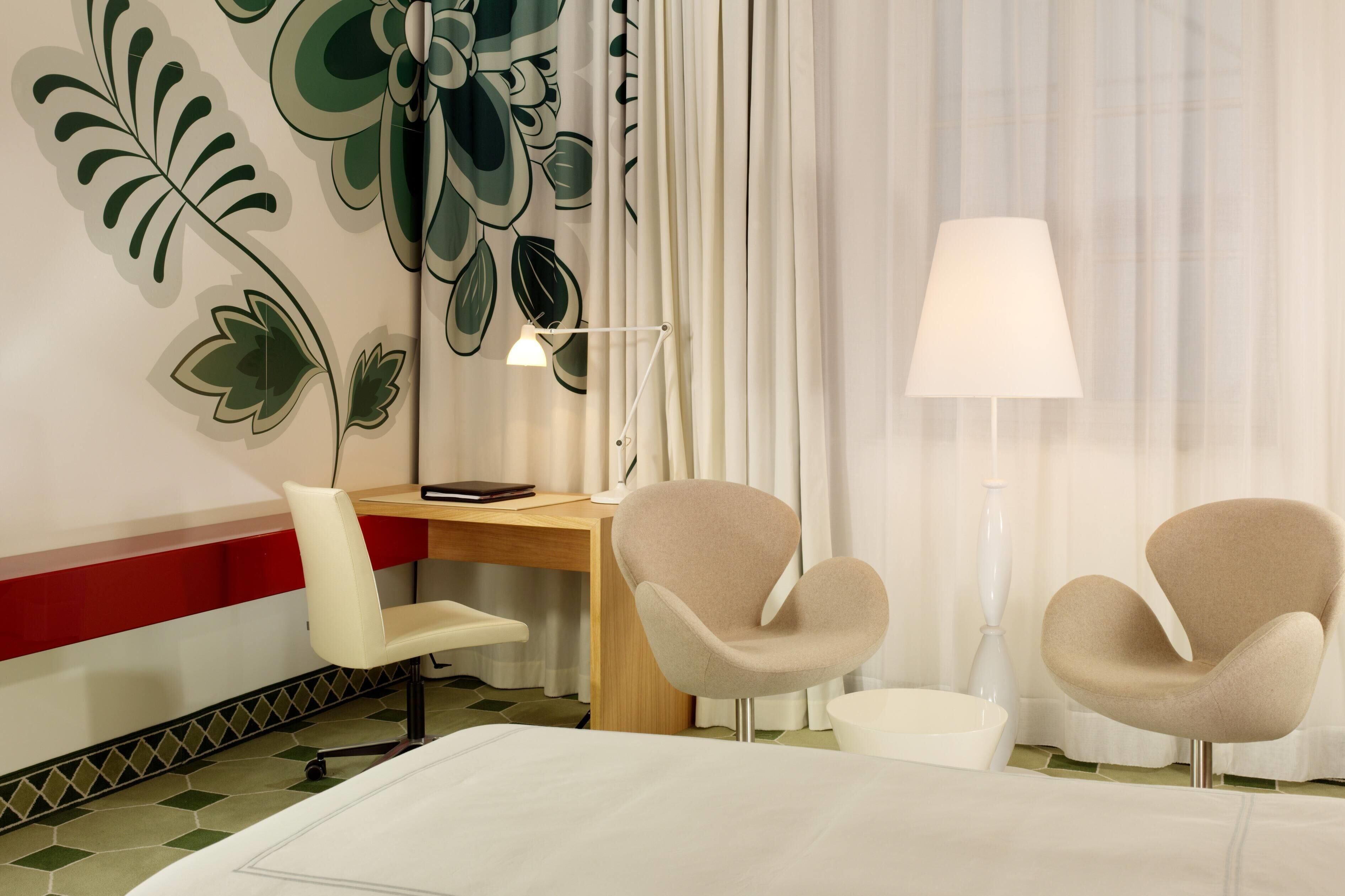 A hotel room with a bed, a desk, two chairs, a lamp, and a wall with a leaf pattern.