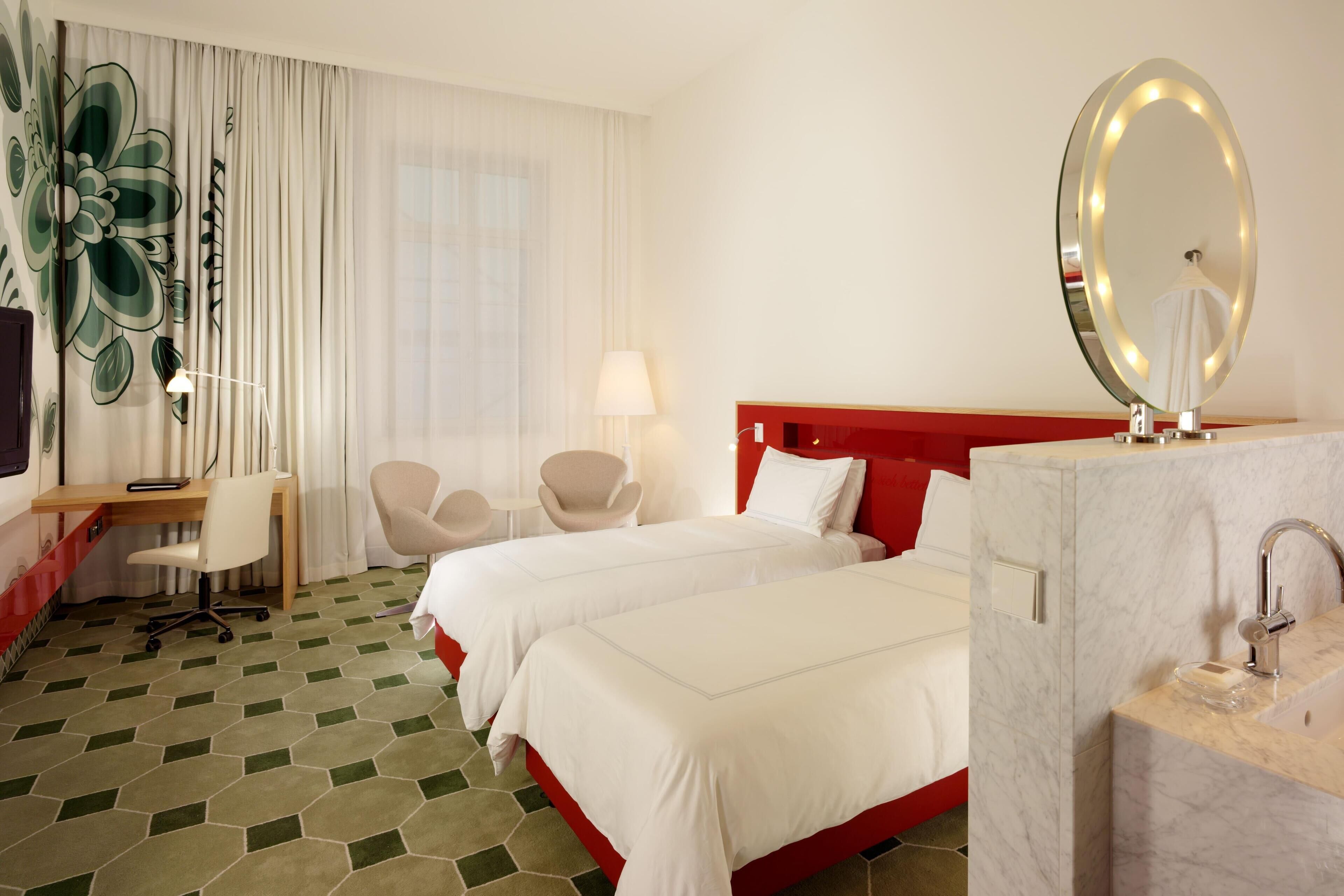 A hotel room with two beds, a desk, a chair, a vanity with a mirror, and a window with curtains.