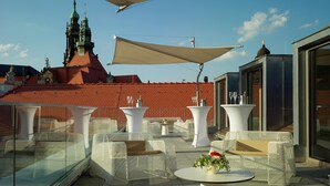 Una terraza en la azotea con mobiliario exterior, una mesa con un florero y vistas a una iglesia de tejado rojo y aguja verde.