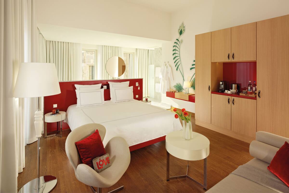Een hotelkamer met een groot bed, een zithoek met een stoel en een tafeltje, een spiegel, een rode accentmuur en een kitchenette.