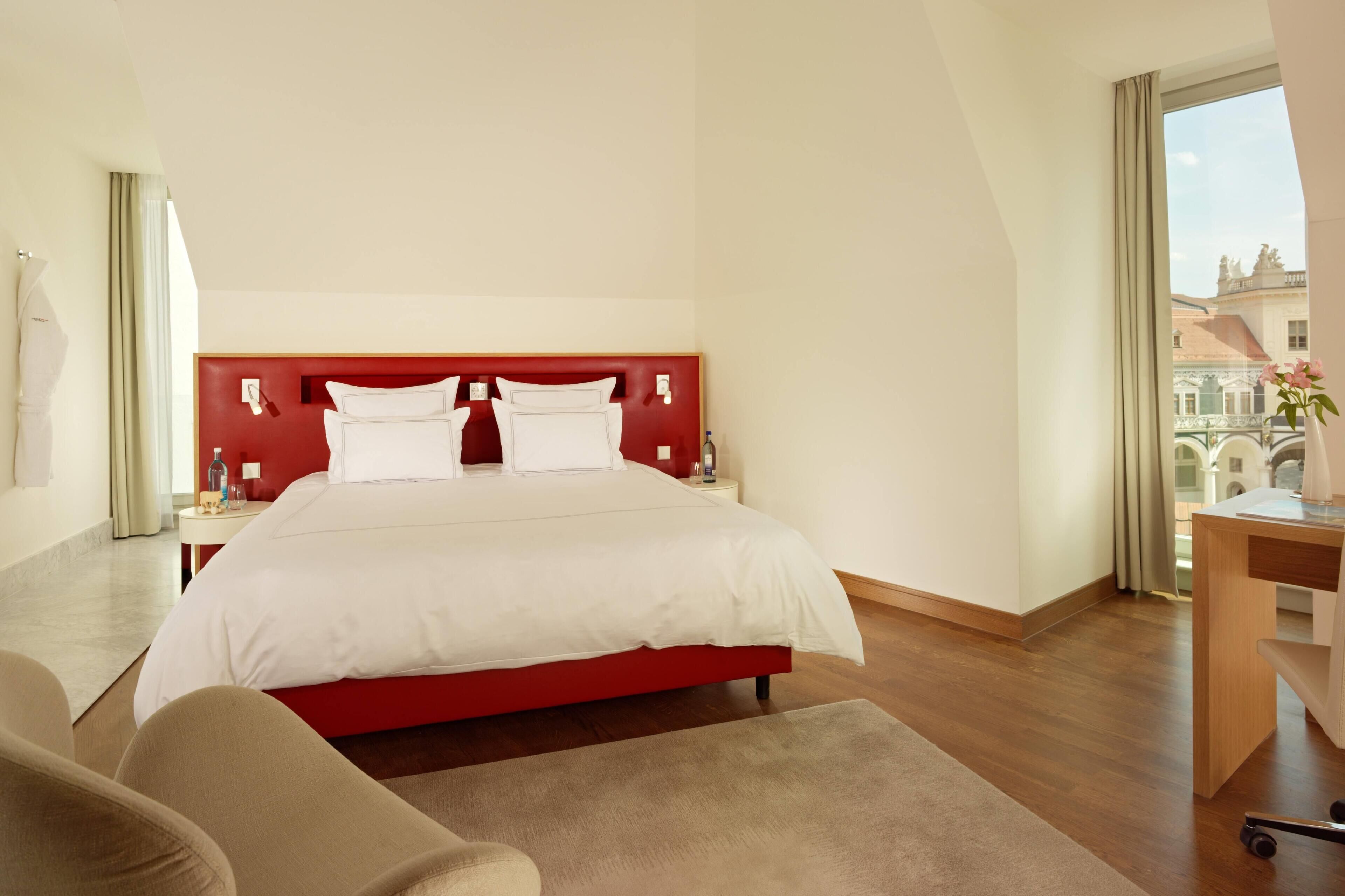 A hotel room with a red headboard, a large bed, a desk, and a view of buildings through the window.
