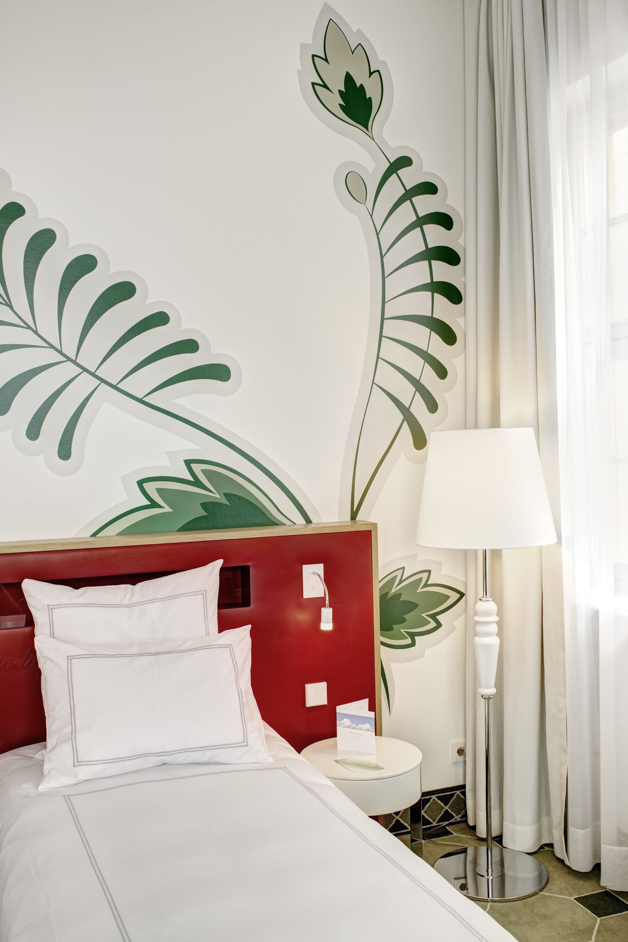 Habitación de hotel con una cama, un cabecero rojo, un cuadro de una hoja, una lámpara blanca y un baño con espejo y toallero.