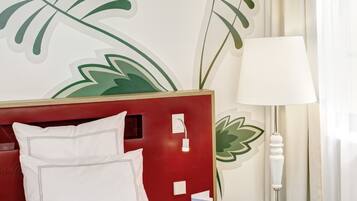 Habitación de hotel con una cama, un cabecero rojo, un cuadro de una hoja, una lámpara blanca y un baño con espejo y toallero.