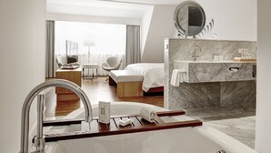 Una habitación de hotel moderna con una cama grande, una zona de estar con una silla y una mesa, un baño con tocador de mármol y un ventanal con cortinas.