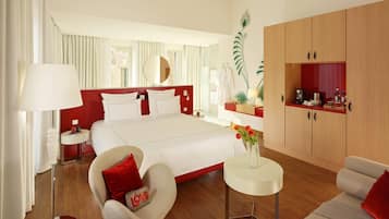 Habitación de hotel con una cama grande, una zona de estar con una silla y una mesita, una kitchenette y un espejo amplio.