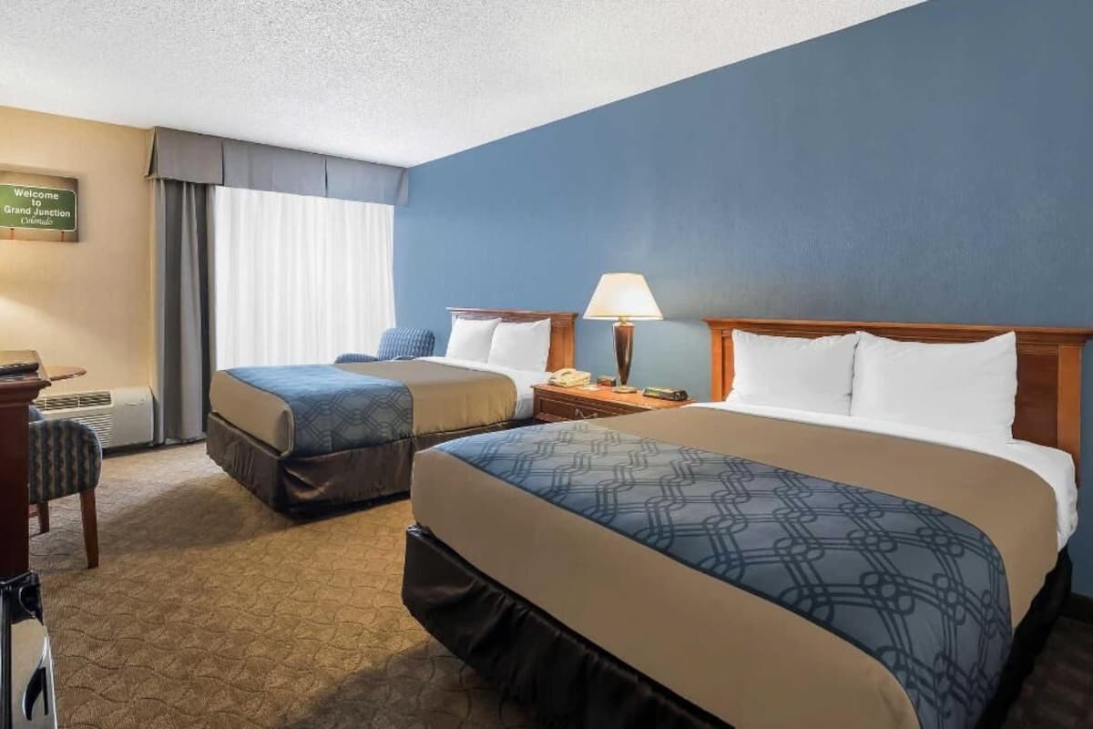 A hotel room with two beds, a desk with a chair, a lamp, and a sign indicating the Grand Junction location.