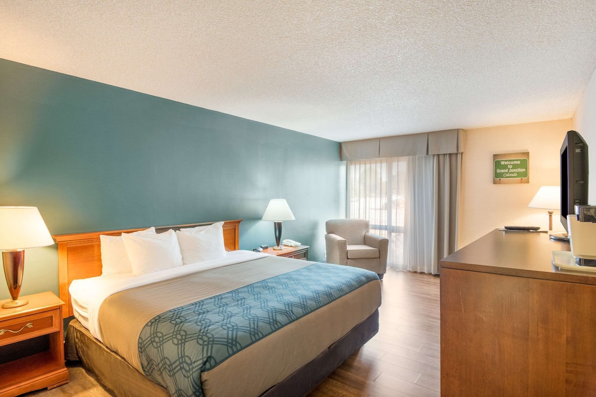 A hotel room with a large bed, two bedside tables with lamps, a chair, a desk with a computer, and a sign indicating the location.
