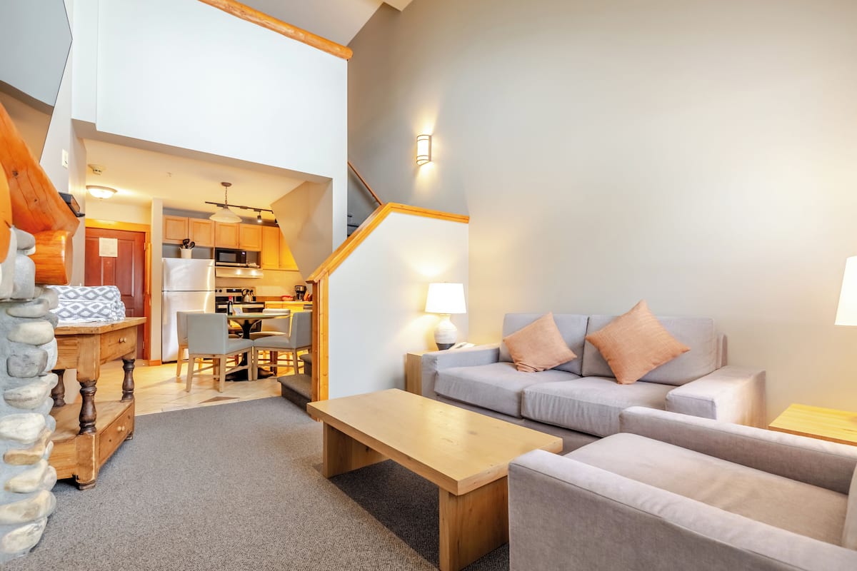 A spacious living area with a sofa, coffee table, and a dining table. A kitchen is visible in the background, and there is a staircase leading to another level.