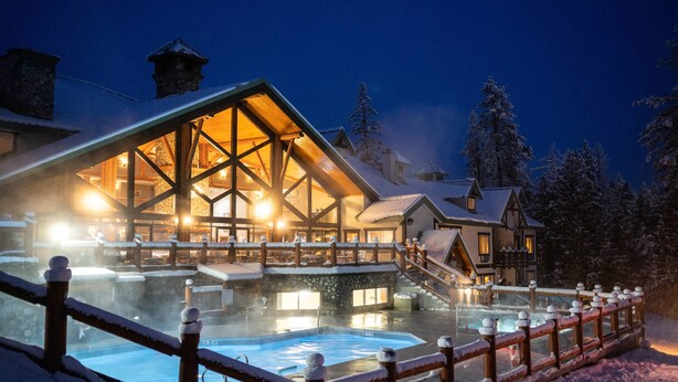 A heated outdoor pool area with a wooden deck and a stone wall, surrounded by snow-covered trees. - Lizard Creek Lodge (Fernie)