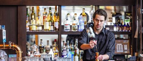 Un barman prépare un cocktail derrière un comptoir, avec différentes bouteilles et une caisse enregistreuse à portée de main.